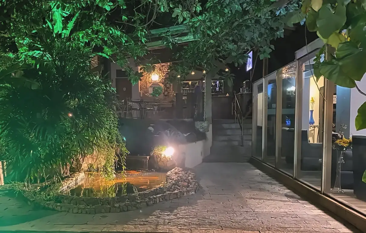 O lago do jardim iluminado à noite, com o caminho de pedra, o bar ao fundo e o lounge de vidro à direita